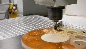 CNC milling bit cutting into a round orange workpiece on a perforated metal worktable, creating circular holes and patterns.