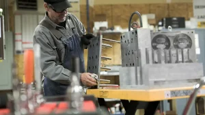 Machinist in safety gear polishing a metal jig with pins on a workbench in a workshop