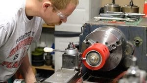 Young man wearing safety glasses leans over a metal lathe, adjusting a workpiece in a workshop.