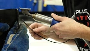 Close-up of a technician's hands guiding a powered hand tool against a metal workpiece, wearing a blue protective glove.