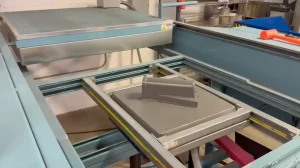 Industrial processing line with blue metal frames and stainless components, showing a conveyor/roller system and an orange-handled tool resting on top.