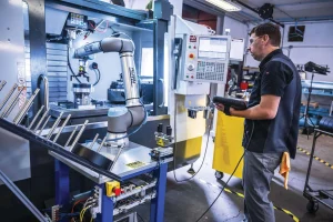 Technician wearing safety glasses and ear protection monitors a robotic machining station with a control panel on screen and machine components nearby.